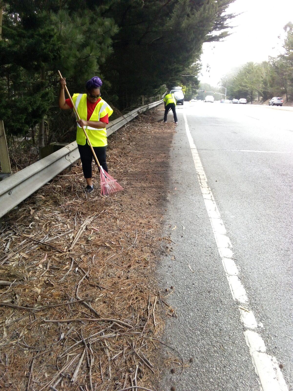 A volunteer in a high visibility vest raking pine duff off the Skyline shoulder into a pile